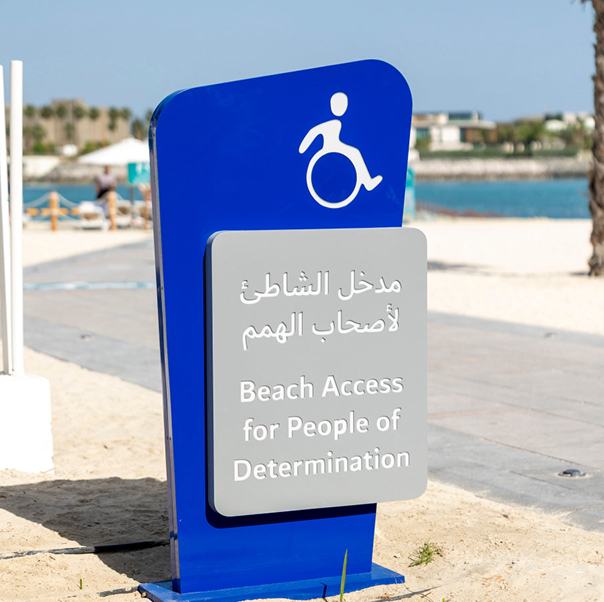 Blue sign with a wheelchair symbol reading Beach Access for People of Determination, in Arabic and English